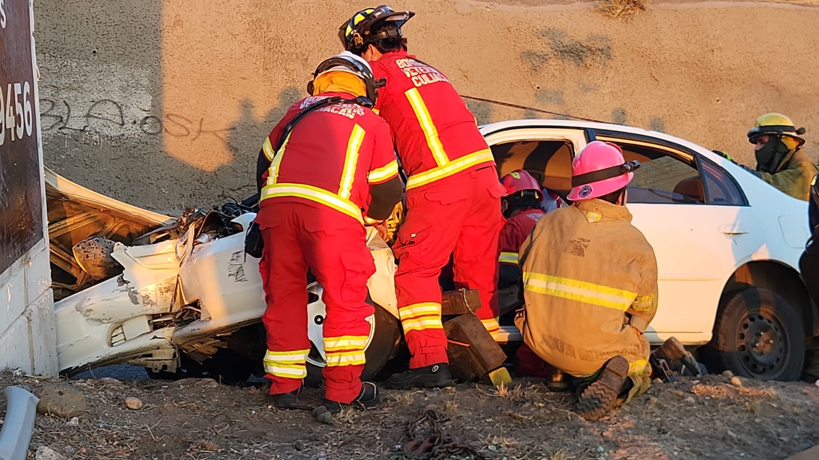 Bomberos junto a un carro blanco chocado contra una barda