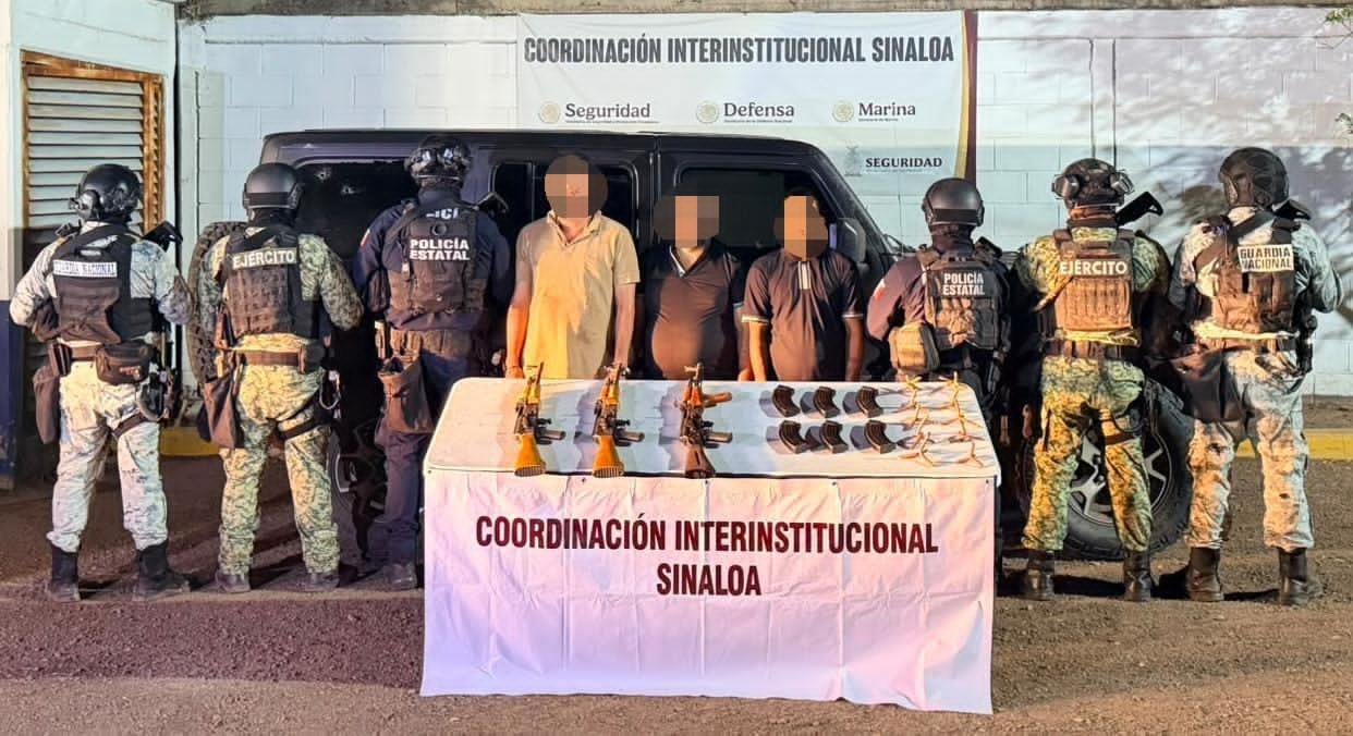 Civiles con el rostro cubierto, a los costados, elementos Militares, al fondo una camioneta negra y al frente una mesa con armas y objetos