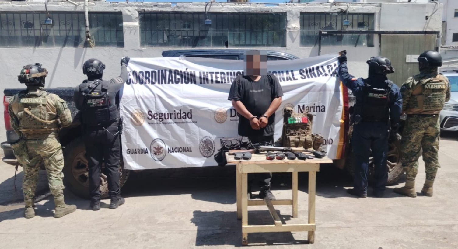 Un hombre detenido junto elementos de seguridad una lona del ejército mexicano detrás