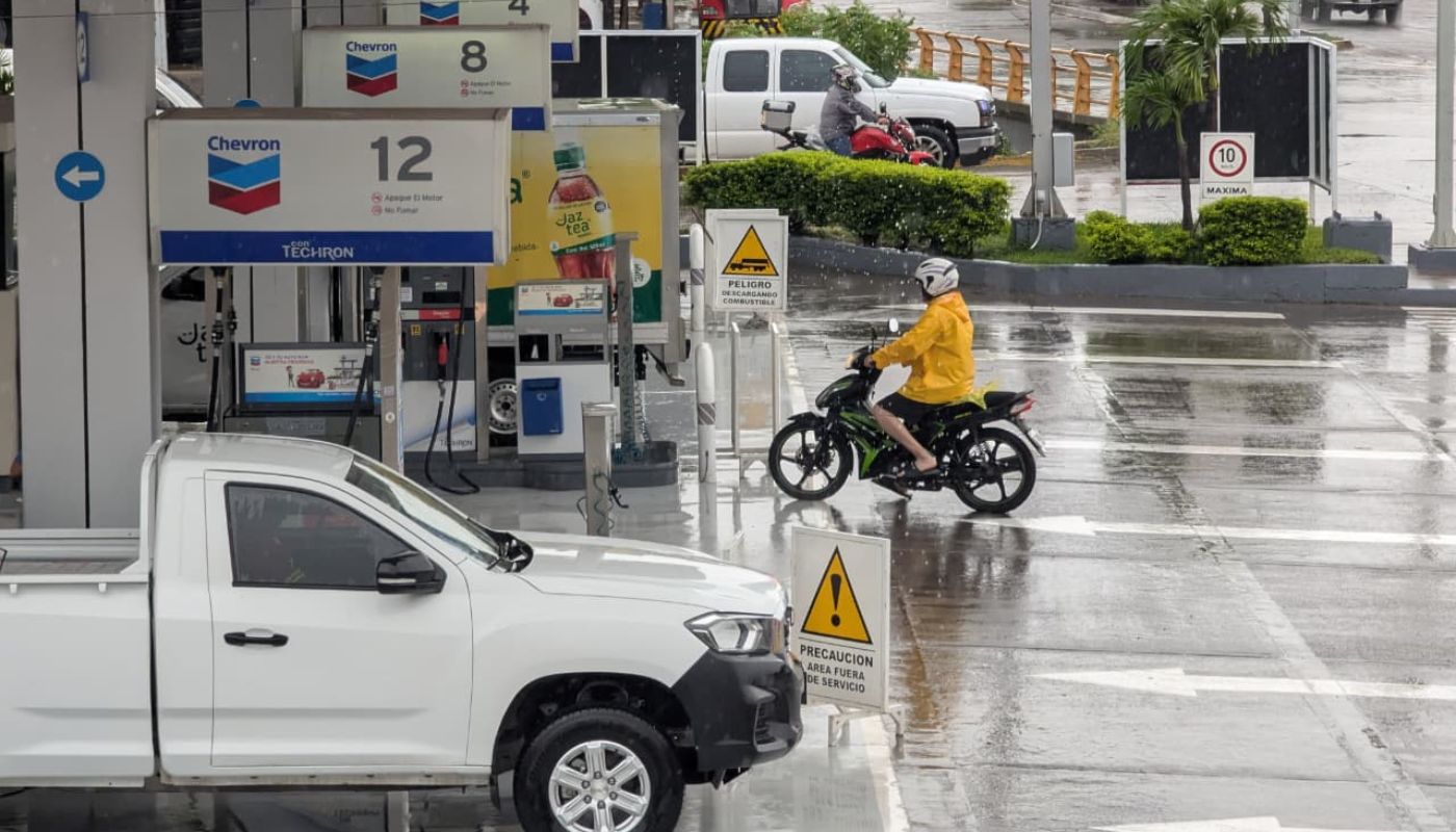 PErsona con impermeable arriba de una moto y una camioneta blanca en una estación de servicios, piso mojado tras la lluvia