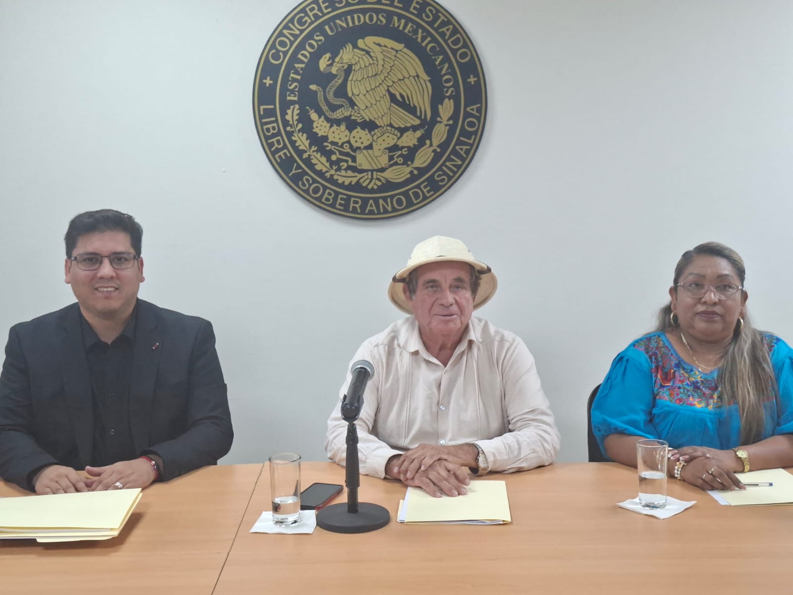 Tres personas con un micrófono y el escudo del congreso de Sinaloa
