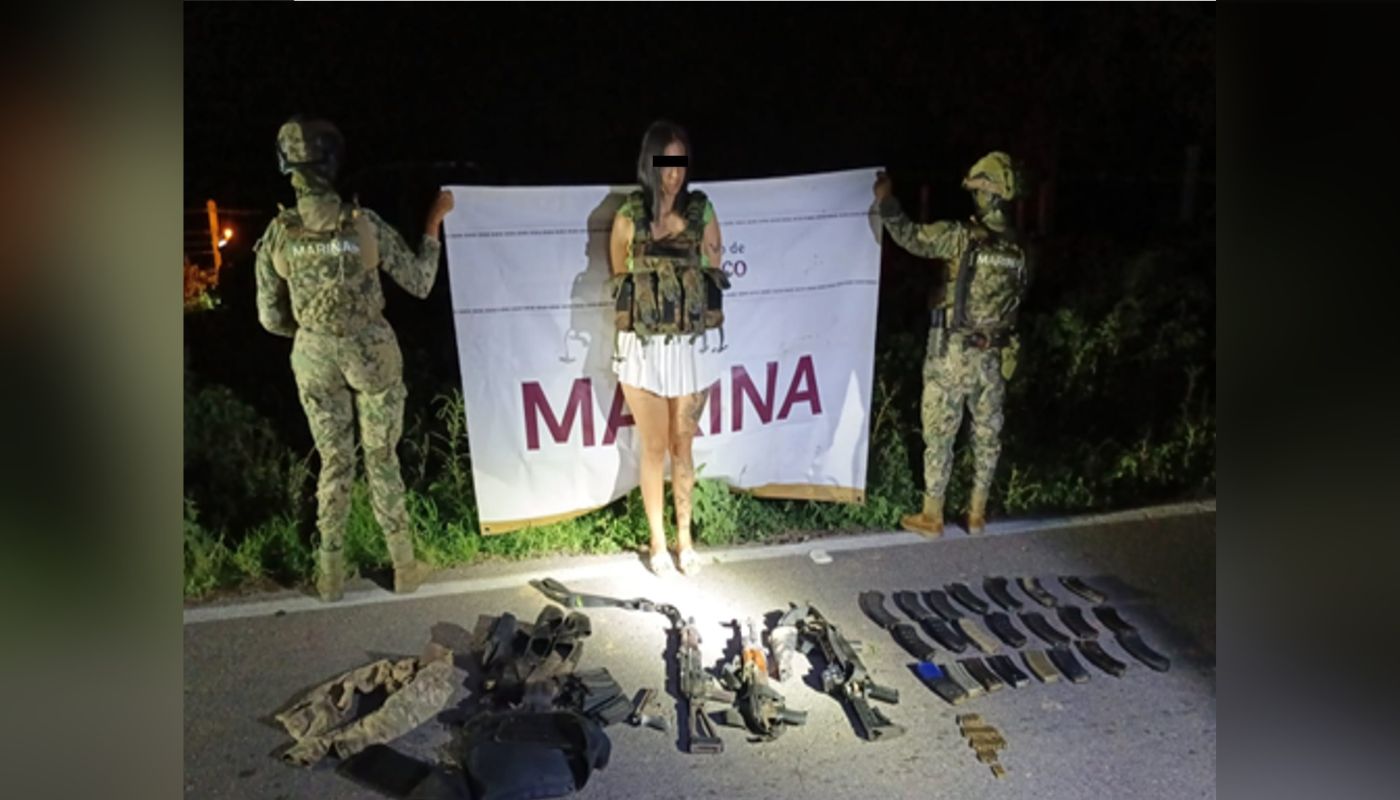 civil con el rostro cubierto, las manos en las espaldas y con un chaleco táctico junto a dos elementos militares que sostienen una lona blanca con la leyenda Marina, en la parte inferior, armas y objetos