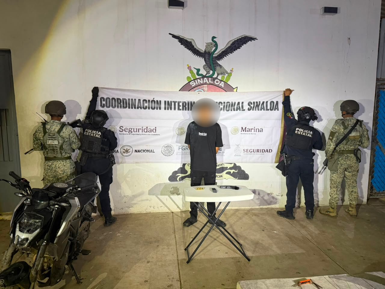 civil con el rostro cubierto y las manos en la espalda, a los costados, cuatro elementos policiacos de espaldas sosteniendo una lona con logos de corporaciones de seguridad, en la parte inferior un arma